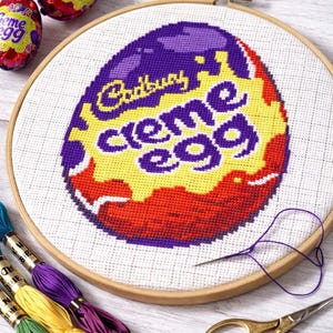 May include: A cross-stitch embroidery of a Cadbury Creme Egg in a wooden hoop. The egg design features purple, yellow, and red colors. Embroidery floss in green, yellow, and purple, along with scissors and a needle, are also visible.