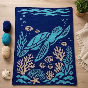 May include: A rectangular crochet blanket featuring a dark blue background with a large light blue sea turtle swimming among beige coral, light blue seaweed, and small fish. The design includes bubbles and wavy lines at the top of the blanket.