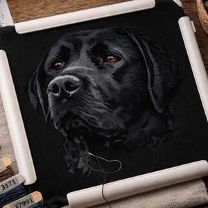 May include: A black Labrador Retriever cross-stitch in a white frame. The dog's portrait is detailed with brown eyes. Embroidery threads and a basket of supplies are on a wooden surface, alongside scissors.