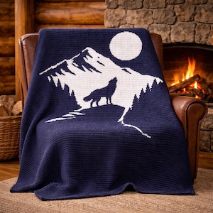 May include: A navy blue knitted throw blanket draped over a leather armchair. The blanket features a white graphic of a mountain range, a full moon, and a howling wolf. The background includes a fireplace with a burning fire and a wooden wall.