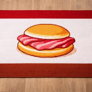 Könnte beinhalten: Rechteckiger Teppich mit einer Cartoon-Illustration eines Burgers. Der Teppich hat eine weiße Mitte, oben rot und unten braun umrandet. Die Burger-Illustration zeigt Brötchen und Speck, zentral platziert.