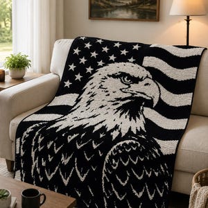 May include: A black and white knitted throw blanket featuring an American flag design with an eagle's head. The blanket has a detailed depiction of the eagle and the flag's stars and stripes. The blanket is draped over a beige sofa.