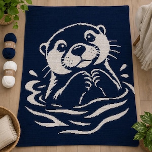 Könnte beinhalten: Eine rechteckige Häkeldecke mit dunkelblauem Hintergrund und einer großen, weißen, detaillierten Grafik eines im Wasser schwimmenden Otters. Der Otter ist vom Oberkörper aufwärts dargestellt, wobei die Pfoten nah am Gesicht gehalten werden.