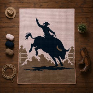 May include: A rectangular crochet blanket featuring a dark navy blue silhouette of a cowboy riding a bucking bull. The background is a light pink, with a brown fence and ground design at the bottom. The blanket has a thin, light brown border.