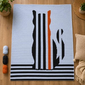 May include: A rectangular crochet blanket featuring a light blue background with a central graphic of a cat silhouette composed of vertical black stripes and one single orange stripe. The bottom edge has horizontal black stripes across the width.