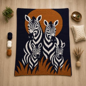 May include: A rectangular crochet blanket featuring a dark navy blue background with a large orange sun. The design displays four zebras in black and white, with two larger zebras standing behind two smaller zebras, all set above orange grass.