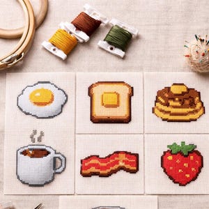 Könnte beinhalten: Kreuzstich-Frühstücksmotive: Ei, Toast, Pfannkuchen, Kaffee, Speck und eine Erdbeere. Das Bild zeigt auch Spulen mit braunem, gelbem und grünem Garn, einen Stickrahmen und ein Nadelkissen.