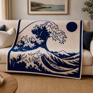 Può includere: Una coperta lavorata a maglia color crema con una raffigurazione blu navy de "La Grande Onda di Kanagawa" di Hokusai. Il disegno include una grande onda, un sole e montagne. La coperta è drappeggiata su un divano beige.