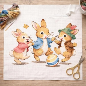 Könnte beinhalten: Kreuzstichprojekt mit drei Cartoon-Kaninchen, die mit einem Ball spielen. Die Kaninchen tragen Jacken und Hüte, mit einem Schmetterling im Hintergrund. Stickgarn, Nadeln und Schere sind ebenfalls sichtbar.