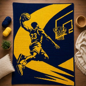 May include: A rectangular crochet blanket featuring a dark blue background with a large yellow graphic of a basketball player wearing a jersey with the number 23, mid-air performing a slam dunk toward a yellow basketball hoop and net design.