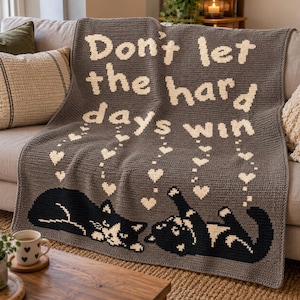 May include: A dark grey crocheted blanket featuring the cream-coloured text "Don't let the hard days win" at the top. Below the text are small cream-coloured hearts and two black and white cats, one lying down and one lying on its back.