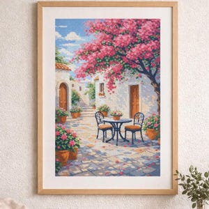 May include: Framed cross-stitch artwork depicting a sunny courtyard scene. A table and chairs sit on a stone patio beneath a blooming pink tree. White buildings with brown doors and flower pots line the courtyard, under a blue sky.