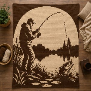 Puede incluir: Una manta de ganchillo rectangular con un diseño bicolor en marrón y crema. El patrón muestra a una persona de pie en la orilla pescando con una caña, con un pez grande saltando fuera del agua en primer plano y árboles al fondo.