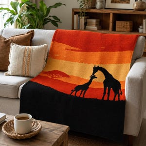 Op de afbeelding: Een gebreide sprei met een zonsondergangscène met silhouetten van giraffen. De sprei heeft een zwarte basis met oranje, gele en rode strepen. De sprei is over een grijze bank gedrapeerd, wat een warme uitstraling creëert. Afmetingen ca. 150 cm x 200 cm.