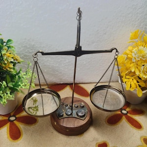 May include: A vintage brass balance scale with two circular pans suspended by chains. The scale has a wooden base with small weights. The background includes artificial flowers and a patterned surface.