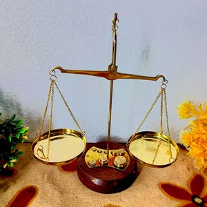 May include: A vintage brass scale with two round pans suspended by chains. The scale has a wooden base with brass accents and a central vertical support. The scale is designed for weighing items and has a classic, antique aesthetic.