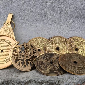 Puede incluir: Una colección de astrolabios antiguos de latón y componentes relacionados. La pieza más grande tiene un asa y texto grabado. También hay varios discos circulares con diseños y texto intrincados. Los artículos se exhiben sobre una superficie texturizada.