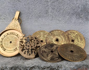 May include: A brass astrolabe featuring a circular main body with an engraved floral handle and a central plate containing text, accompanied by five separate circular brass plates with intricate etched celestial maps and navigational markings.