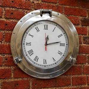 Könnte beinhalten: Eine runde, silberfarbene Wanduhr mit weißem Zifferblatt und schwarzen römischen Ziffern. Die Uhr hat schwarze Zeiger und einen roten Sekundenzeiger. Sie ist an einer roten Backsteinmauer befestigt. Das Design der Uhr ähnelt einem Schiffsbullauge.