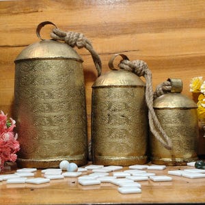 May include: Three gold-colored metal bells with intricate embossed patterns. Each bell has a rope handle. The bells vary in size, with the largest bell measuring approximately 10 inches tall. The bells are displayed on a wooden surface.