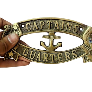 Pode incluir: Uma placa oval de latão com o texto CAPTAINS QUARTERS em relevo. O centro exibe um ícone de âncora náutica, enquanto as laterais são decoradas com motivos de águias e estrelas em relevo sobre um fundo texturizado com furos de montagem.