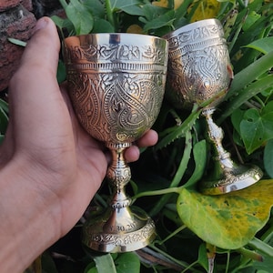 May include: Two ornate brass goblets with intricate floral and scrollwork designs. The goblets have a pedestal base and are held in a hand against a backdrop of green foliage. The date "3.30.20" is engraved on the base of one goblet.