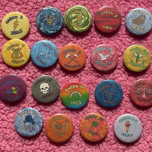 Könnte beinhalten: Eine Sammlung bunter runder Knöpfe mit verschiedenen Designs und Texten. Jeder Knopf zeigt ein anderes Bild und das Wort "Cabin" gefolgt von einer Nummer. Einige Knöpfe haben Bilder und Namen im Zusammenhang mit griechischen Göttern. Die Knöpfe liegen auf einer rosa, strukturierten Oberfläche.