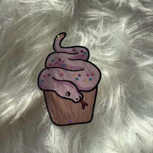May include: A sticker depicting a cupcake with a snake design. The snake is wrapped around the light purple frosting, which has colourful star sprinkles. The cupcake base is brown, and the snake has a red tongue.