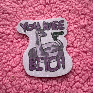 Peut inclure: Autocollant avec une créature violette ressemblant à un dinosaure tenant une arme à feu. Le texte "You Wee Bitch" est écrit en lettres violettes. L'autocollant a un fond violet clair avec des étoiles blanches.