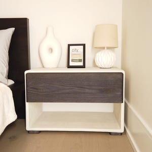 May include: A modern nightstand with a dark wood drawer and a white frame. The nightstand has a white vase, a framed picture with the word "city", and a white lamp with a beige lampshade. The nightstand is placed next to a bed.