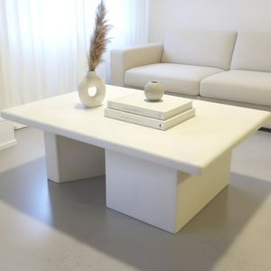 May include: A modern, rectangular white coffee table with a minimalist design. The table features a solid top and two rectangular supports. Decorative items on the table include a vase with a circular opening, a small round vase, and two books. A beige sofa is in the background.