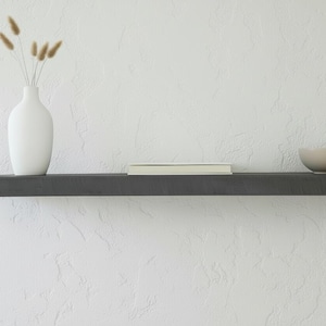 May include: A dark grey floating shelf against a white textured wall. The shelf holds a white vase with dried flowers, a closed book, and a small grey bowl. The shelf is a simple, modern design.