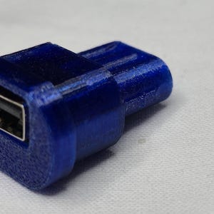 Könnte beinhalten: Ein blauer USB-Adapter mit rechteckiger Öffnung. Der Adapter besteht aus einem strukturierten Material und ist für den Anschluss an einen USB-Anschluss konzipiert. Das Bild ist auf einer weißen Oberfläche aufgenommen.