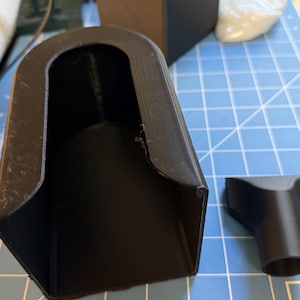 May include: A black 3D-printed plastic component featuring a U-shaped opening and a smooth interior surface. The object has a matte finish and is positioned alongside a smaller, separate black plastic attachment with a tapered cylindrical end.