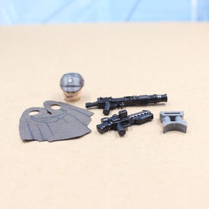 May include: A collection of miniature toy accessories, including a silver helmet, a gray cape, two black rifles, and a gray block. The items are likely for a miniature figure or toy set.
