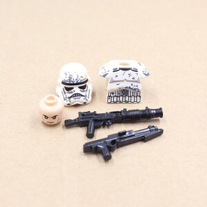 May include: A collection of miniature toy figures and weapons. Includes a white and black Stormtrooper helmet, a tan head, a white and gray torso, and two black toy guns. The items are arranged on a light brown surface.