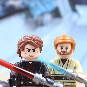 May include: Two Lego minifigures, one with a red lightsaber and the other with a blue lightsaber. The minifigure with the red lightsaber has brown hair and a stern expression. The other minifigure has orange hair and a beard.