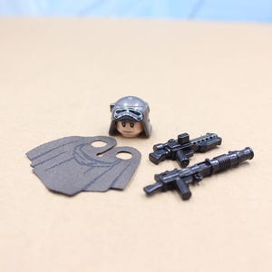 May include: A collection of miniature toy accessories, including a helmet, a cape, and two black rifles. The helmet is silver and brown, and the cape is gray. The rifles are detailed with various components.