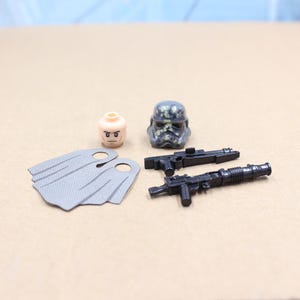 May include: A collection of miniature toy accessories, including a Stormtrooper helmet with camouflage detailing, two black blasters, a light gray cape, and a minifigure head with a neutral expression.