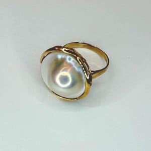 May include: A gold ring featuring a large, lustrous, off-white pearl. The pearl has iridescent highlights and is set in a gold bezel. The ring's band is also gold, creating a classic and elegant design.