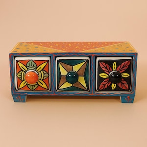 Puede incluir: Una caja de almacenamiento de madera pintada a mano con tres cajones. Cada cajón presenta un diseño floral único y un pomo colorido. La caja está pintada en tonos de azul, amarillo, naranja y verde, con un patrón decorativo en la parte superior.