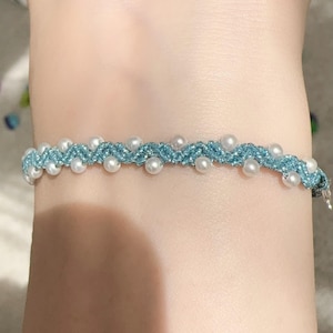 Könnte beinhalten: Ein zartes Armband mit einem gewebten hellblauen Band, das mit kleinen, gleichmäßig verteilten weißen Perlen verziert ist. Das Armband wird mit einem kleinen Verschluss befestigt. Dieses Schmuckstück ist für den Alltag geeignet.
