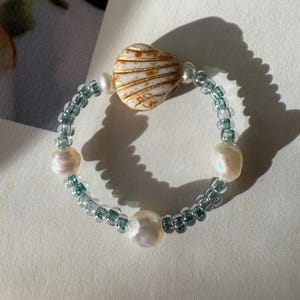 May include: A beaded ring featuring a central white and gold shell-shaped bead, interspersed with translucent teal glass seed beads and three irregular white freshwater pearls arranged symmetrically around the circular band.