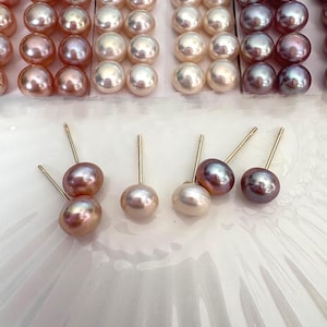 May include: Rows of lustrous, round pearls in shades of pink, white, and gray. Several pairs of pearl stud earrings with gold posts are displayed, showcasing the variety of colors and the jewelry's elegant design. The pearls have a smooth, reflective surface.