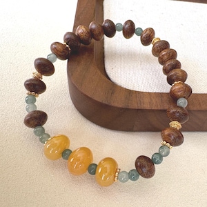 May include: A beaded bracelet featuring dark brown wooden beads, light green beads, and three yellow-orange beads. The bracelet has gold-colored accents and is displayed on a wooden stand. The bracelet is a piece of jewelry.