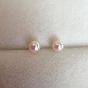 May include: Two small, round, off-white pearls with a subtle iridescent sheen. The pearls are set against a soft, light-colored background, suggesting a jewelry presentation or storage setting. The pearls are a classic jewelry item.