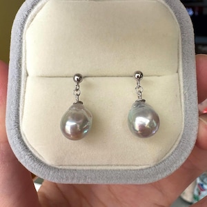 May include: A pair of pearl drop earrings displayed in a gray jewelry box. The earrings feature a silver post and a large, iridescent, teardrop-shaped pearl. The pearls have a lustrous, silvery-gray color with hints of pink and green.
