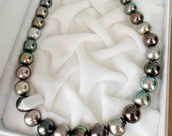 May include: A necklace of iridescent, dark-colored pearls. The pearls range in color from silver and gray to black and bronze, with hints of green and pink. The necklace is displayed in a white, fabric-lined box, suggesting a jewelry presentation.