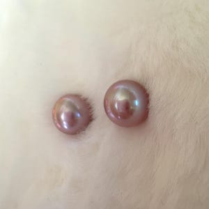 May include: Two lustrous, round, mauve-colored pearls. The pearls have a smooth, iridescent surface, reflecting hints of gold and green. They are set against a soft, white, textured background, creating a delicate and elegant aesthetic. These pearls are ideal for jewelry making.