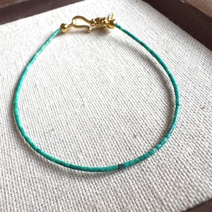 Pode incluir: Uma delicada pulseira de contas turquesa com fecho dourado. A pulseira é feita de pequenas contas turquesas retangulares enfiadas. O fecho é do tipo gancho e olho. A pulseira é exibida em tecido de cor neutra.